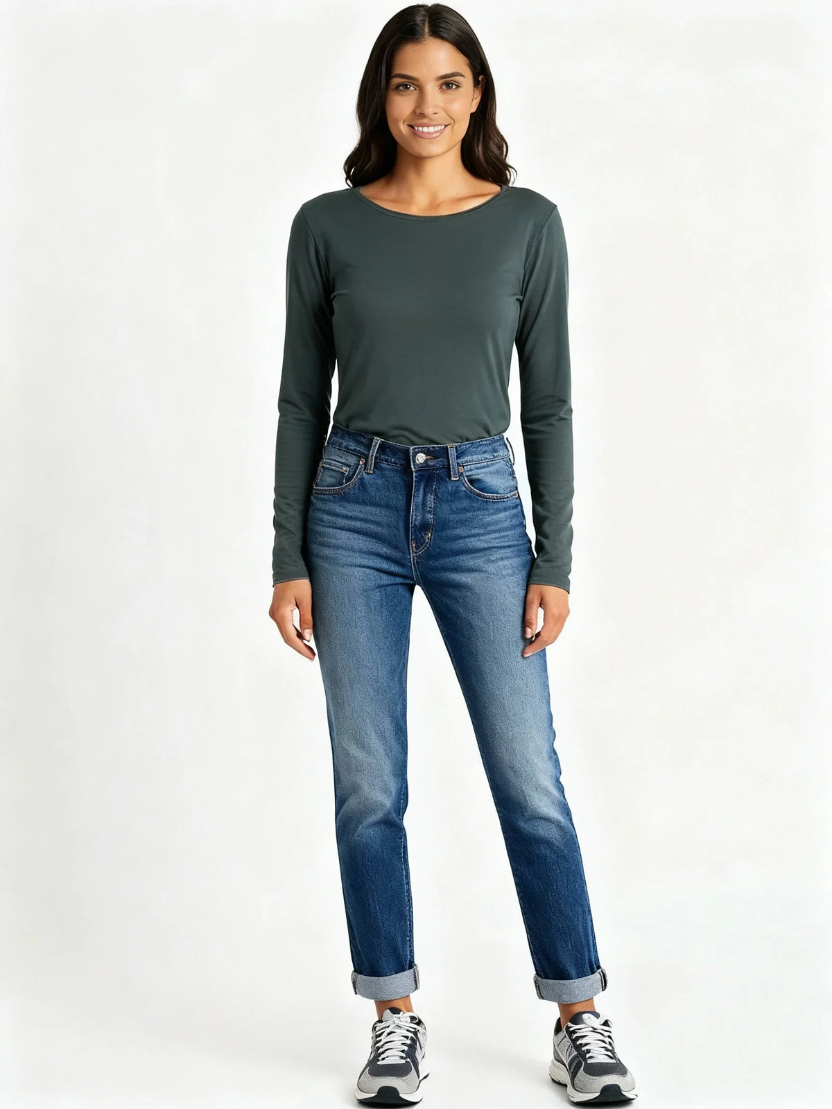 Smiling woman wearing spruce green long sleeve top, cuffed blue jeans, and black white sneakers