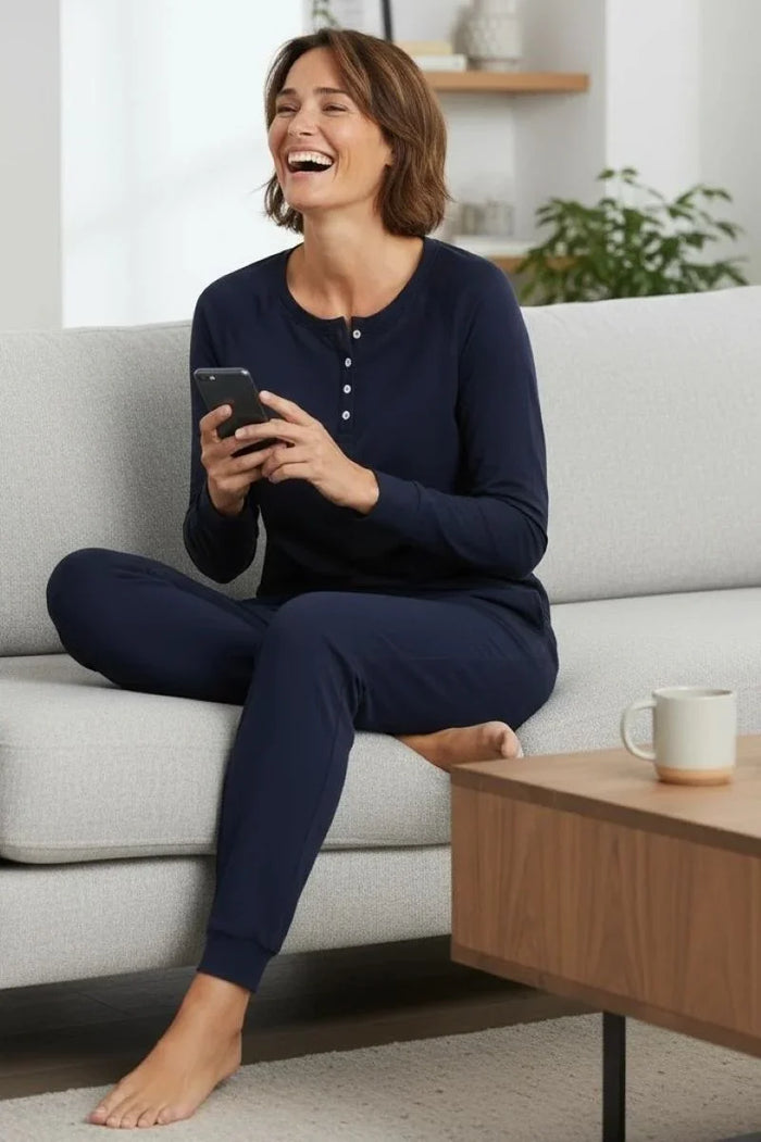 Smiling woman in midnight blue lounge wear sitting cross-legged on gray sofa holding smartphone